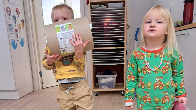 Kaksi lasta päiväkodissa katsomassa kameraan päin, toisella sylissään lelupakkaus.