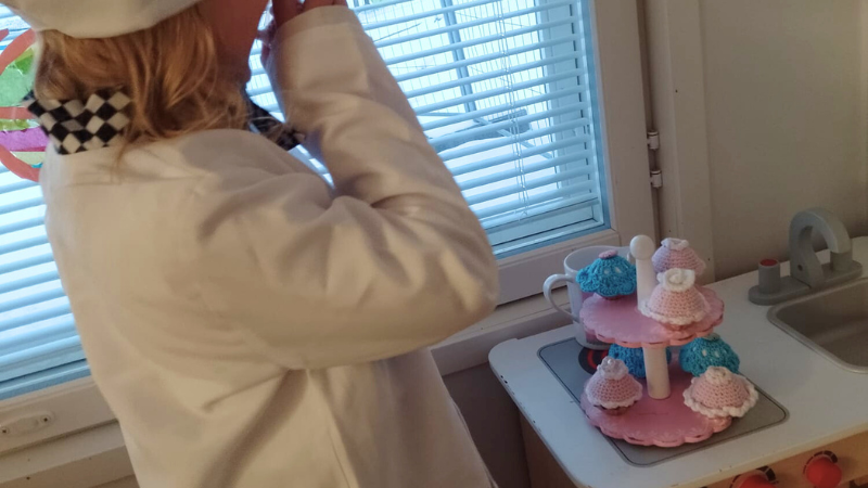 Kokkivaatteisiin pukeutunut lapsi leikkikeittiössä virkattujen kuppikakkujen kanssa.