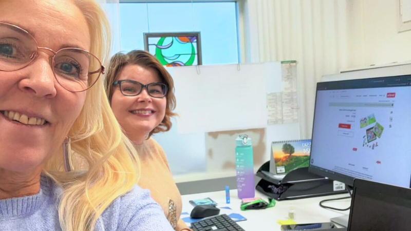 Kaksi naista poseeraamassa selfiessä tietokoneen näytön edessä, jossa meneillään lelutilaus.