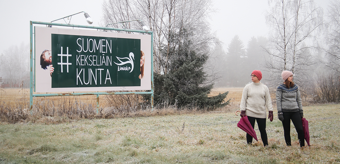 Kaksi naista seisoo #SuomenKekseliäinKunta-kyltin edessä pellolla.
