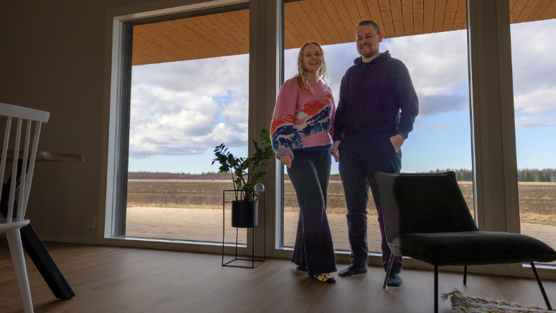 Iida ja Ilari Kinnunen seisovat koko seinän korkuisen ikkunan edessä olohuoneessaan. Taustalla näkyy keväinen peltomaisema.