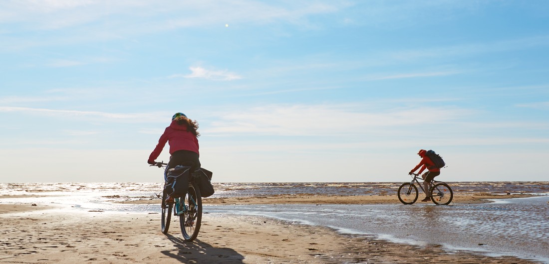 Kaksi pyöräilijää pyöräilee hiekkarannalla aurinkoisessa säässä.