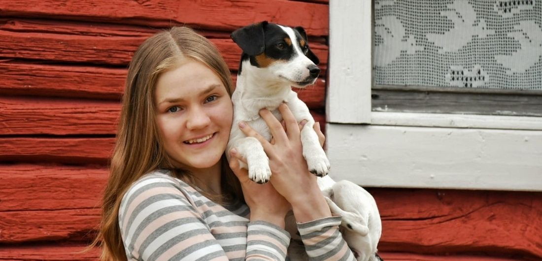 Maija Tuomala koiransa kanssa, taustalla punainen hirsirakennus.