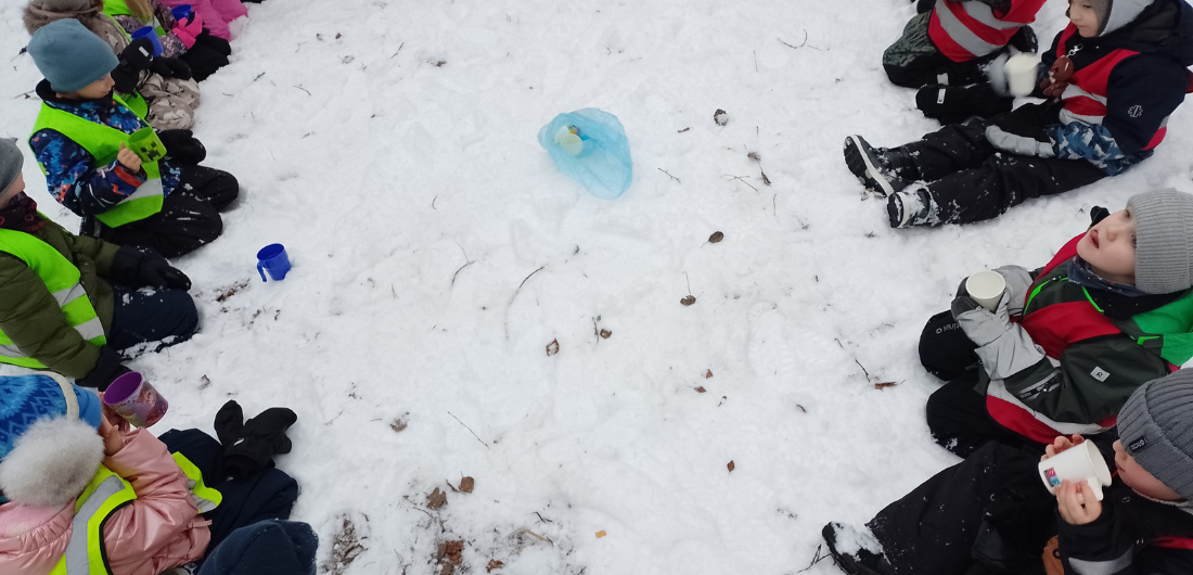 Lapset istuvat ringissä lumihangella ja juovat mehua.