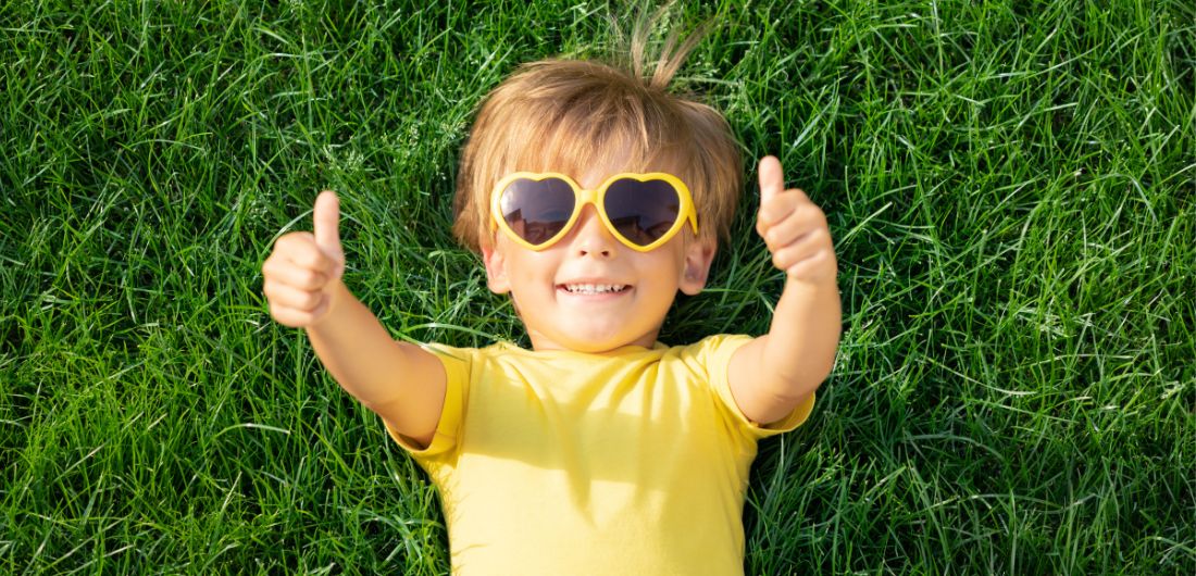 lapsi makaa nurmikolla aurinkolasti päässä ja nostaa peukkua