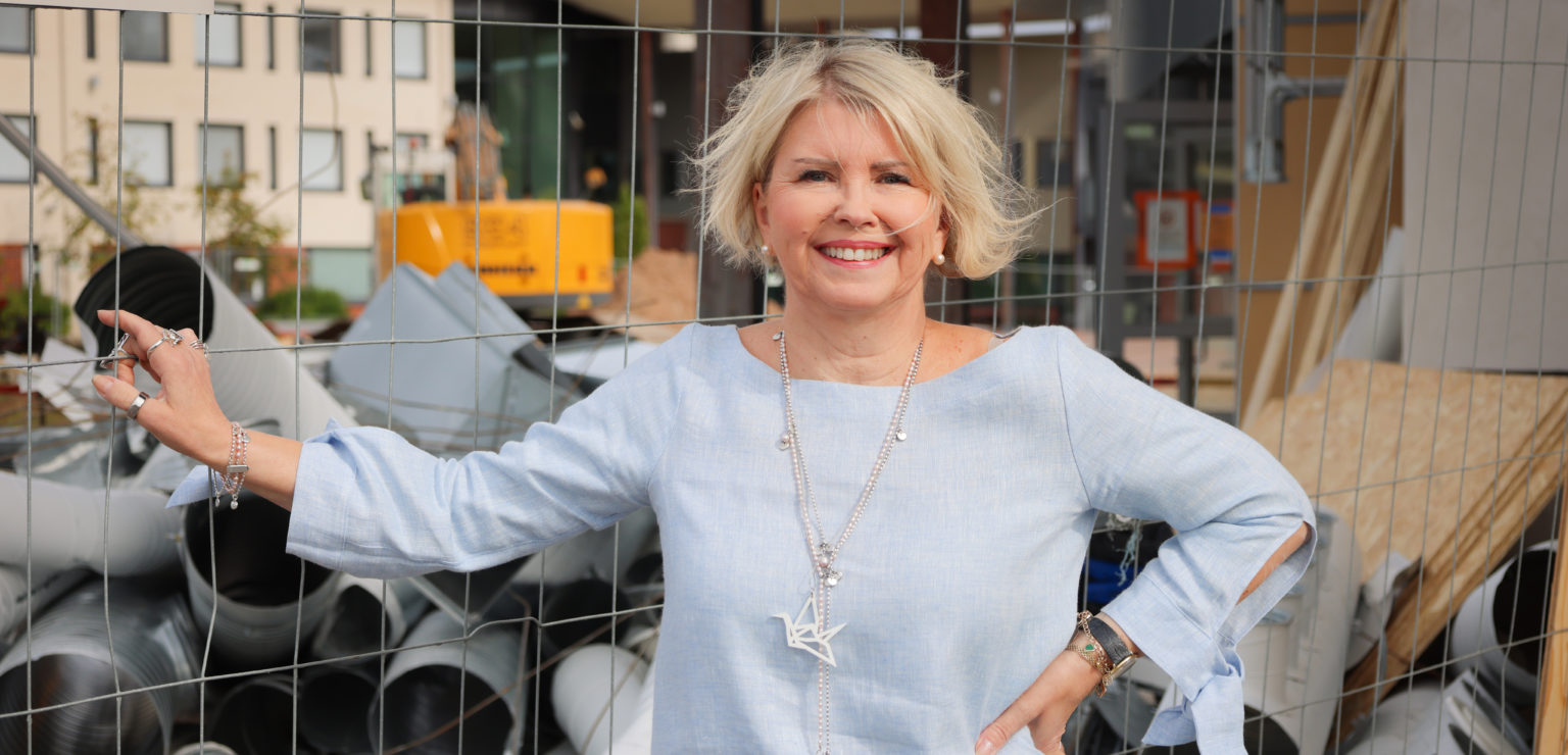 Sivistysjohtaja Päivi Mäki seisoo saneerattavan lukion rakennustyömaa-aidan edessä ja hymyilee.