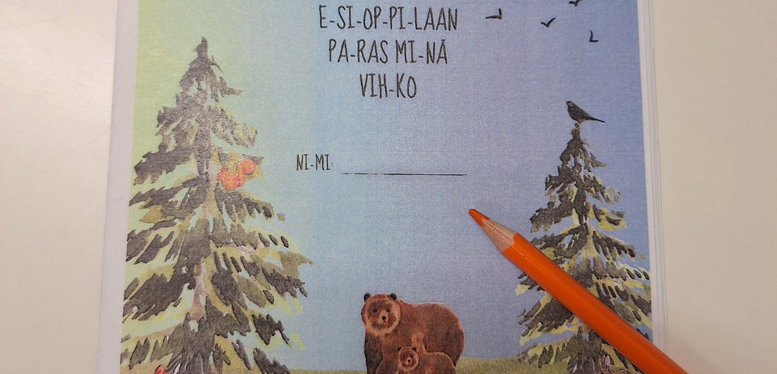 Esioppilaan paras minä -vihko pöydällä oranssin puukynän kanssa. Vihkon kannessa piirrettynä lumisia puita ja karhu pentuineen.