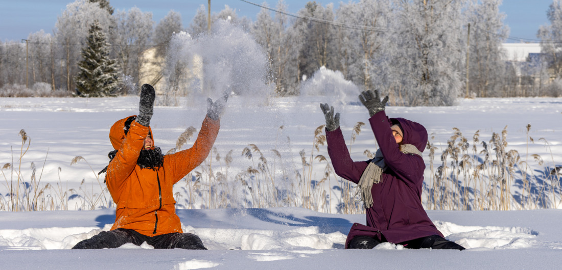 Kaksi naista istuu aurinkoisessa säässä talvivaatteissa lumihangessa ja heittää lunta päänsä yläpuolelle.