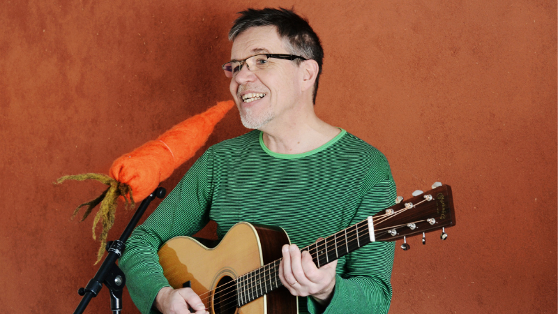 Pentti Rasinkangas soittaa kitaraa ja laulaa porkkanan näköiseen mikrofoniin.