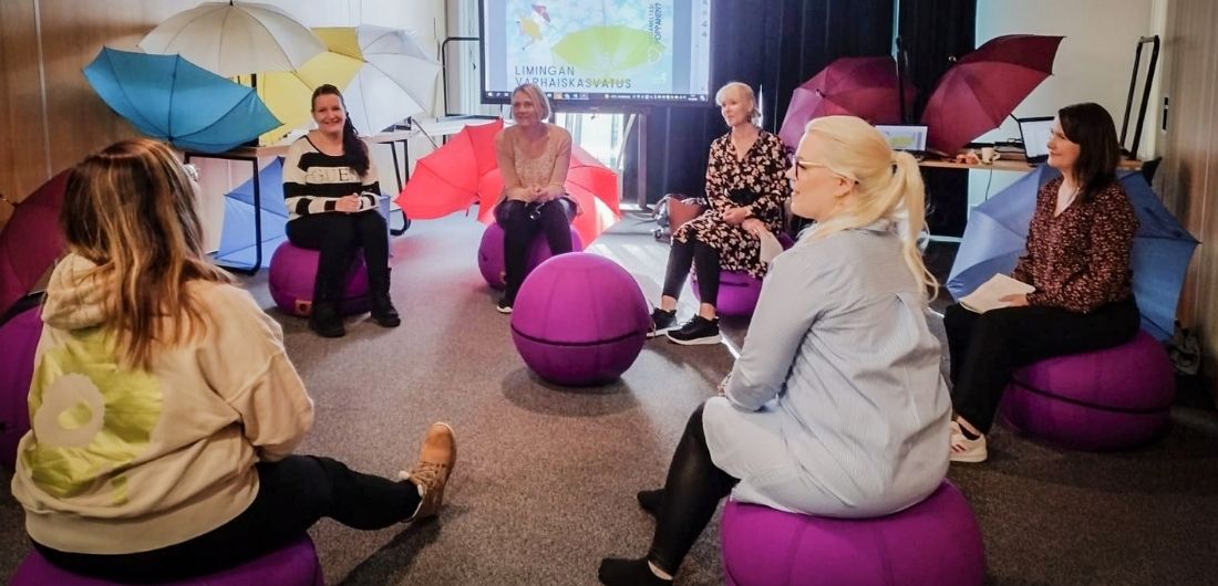 Ryhmä henkilöitä istuu ringissä pallorahien päällä. Tilassa myös värikkäitä koristesateenvarjoja.