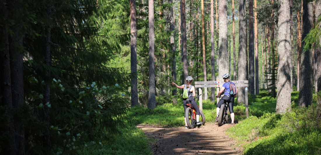 Kaksi pyöräilijää pyöräilemässä maastopyöräilyreitillä ja pysähtyneenä reittimerkkien eteen.