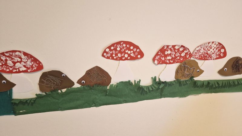 Lasten askartelemia siilejä, myrkkysieniä ja nurmikkoa valkoisella seinällä.