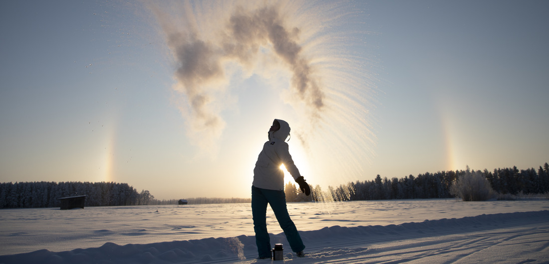 Henkilö on heittänyt vettä ilmaan pakkassäässä ja jäätynyt vesi muodostaa kaaren  ilmaan. Aurinko paistaa henkilön takana.