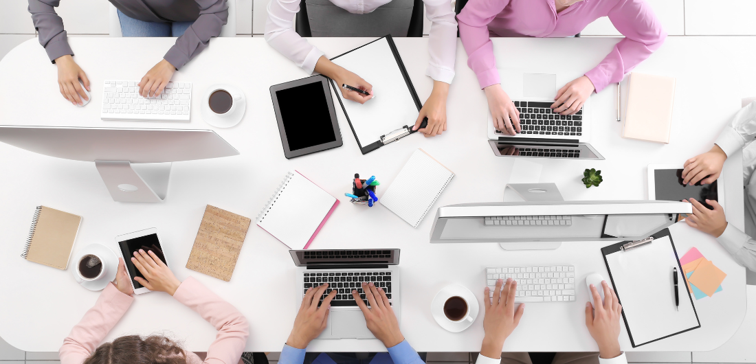 Pöytä kuvattuna ylhäältä, jossa näkyy tietokoneita ja ihmisiä työskentelemässä.