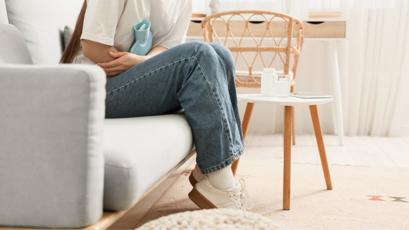 Nainen istuu sohvalla kyyryssä ja pitelee lämminvesipulloa vatsaansa vasten.