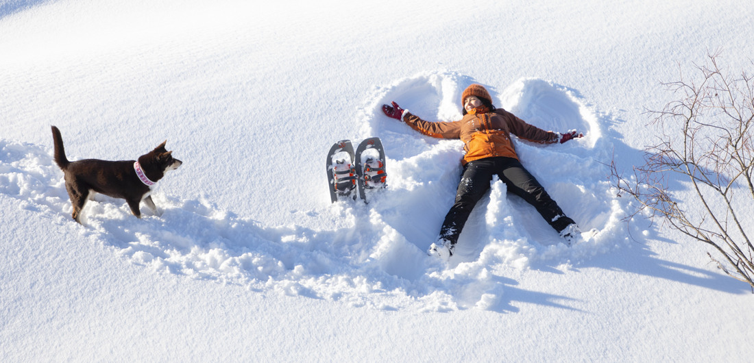 Nainen tekee lumienkelin ja koira katselee vieressä.