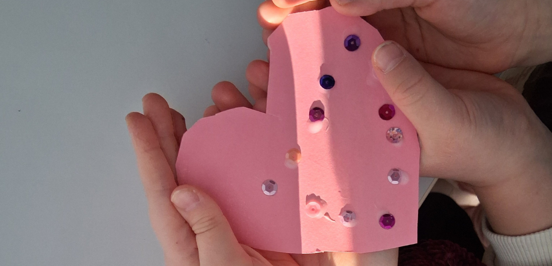 Kädet pitelevät paperista leikattua vaaleanpunaista sydäntä, johon on liimattu koristepaljetteja.