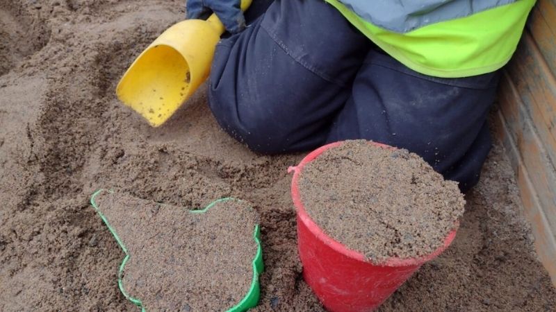 Leikkiämpäri ja hiekkakakkumuotti täynnä hiekkaa ja hiekkalapio lapsen kädessä.