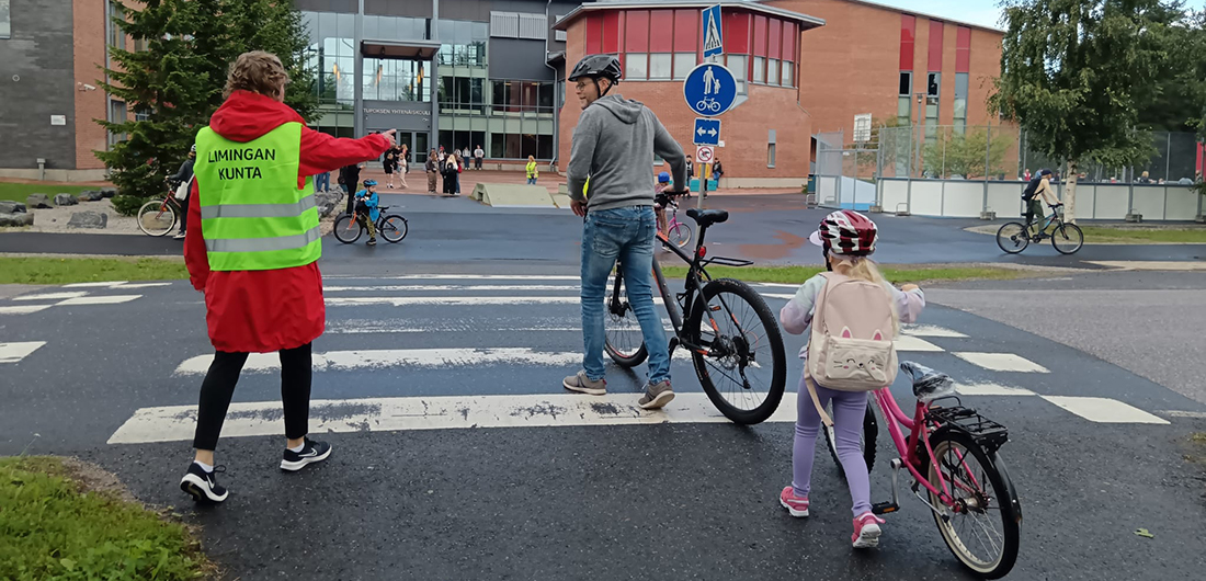 Suojatievahti seisoo suojatien vieressä, isä ja lapsi taluttavat pyöriään.